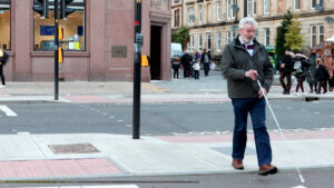 A man with a vision impairment using a long white cane crosses the cycle lane continuing the line of travel from crossing the road. He is unaware the tactile paving for crossing the cycle lane is to his right.